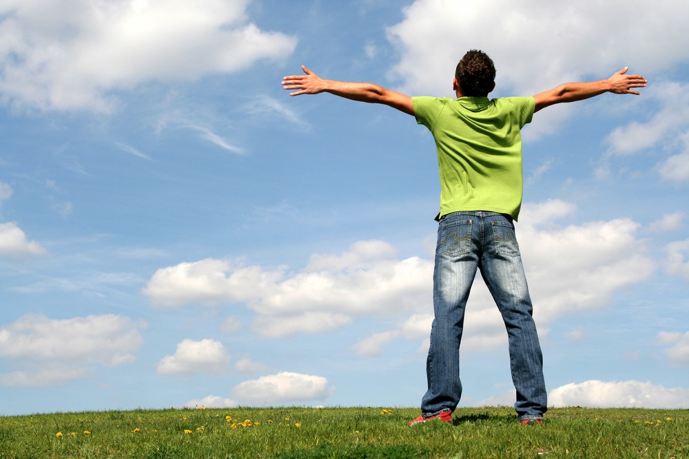 Person with arms spread wide in open field under blue sky