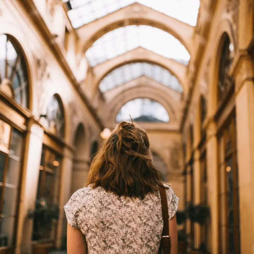 Woman walking forward alone through an archway-lined corridor, viewed from behind.
