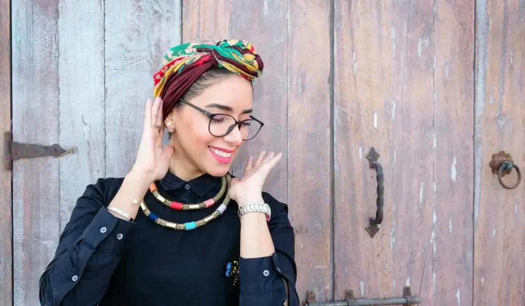 Smiling woman in glasses and colorful headwrap stands confidently against a rustic wooden door.