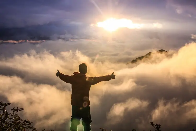 Person standing above the clouds at sunrise with arms raised and thumbs up, symbolizing hope in recovery.